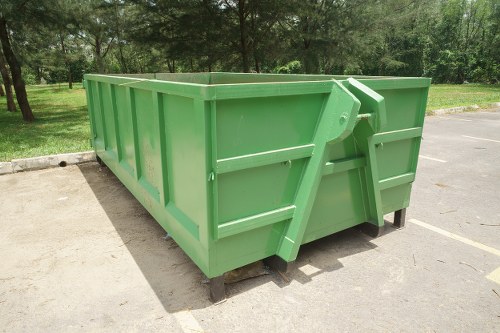 A large, rectangular, green industrial waste skip with vertical ribbing along its sides, positioned on a paved surface in an outdoor area. The skip appears to be made of metal with a smooth, painted finish and is supported on small black wheels or pads at the base. It is situated adjacent to a curb, with a natural, grassy landscape and trees visible in the background, indicating a park or open space setting. The surrounding environment includes a paved parking or driveway area without other visible objects nearby. The lighting is natural, providing clear visibility of the skip's vibrant green colour and structural details. This image is relevant to commercial waste removal services, such as those offered by Commercial Waste Twickenham, illustrating the type of large capacity containers used for collecting and transporting rubbish, suitable for construction, renovation, or business waste management in the Twickenham postcode area.