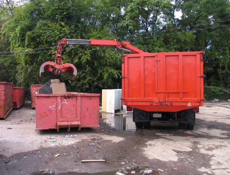 In this outdoor scene, a red waste collection truck equipped with a hydraulic arm and grab attachment is in the process of emptying a large, rectangular red skip positioned on an uneven paved surface. The skip, made of metal with a smooth finish, shows signs of previous use with some minor scuffs and dirt marks on its surface. The hydraulic arm extends over the skip, gripping its top edge firmly as it lifts or tips the container to load waste materials into the truck. Surrounding the main equipment are a few other waste bins of similar size and color, placed on the gravel or asphalt ground that has small patches of dirt and scattered debris, typical of a waste management or industrial area. In the background, a lush green backdrop of trees and foliage indicates a semi-urban environment, possibly near Twickenham or its outskirts. The scene suggests routine rubbish collection work by Commercial Waste Twickenham, focusing on the removal of general waste or construction debris from commercial or industrial premises within the local postcode area, under bright daylight conditions.