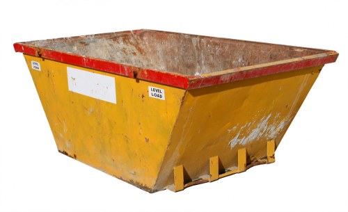 A large industrial skip positioned outdoors on a paved surface, used for commercial waste collection in Twickenham. The skip has a rectangular shape with a wide, open top and sloped sides, constructed from sturdy metal with a bright yellow finish that shows signs of wear and rust. The interior of the skip is visible, revealing a rough, textured surface consistent with heavy-duty usage. The front-facing side displays three small, protruding yellow feet used to support the skip when placed on the ground. Behind the skip, the environment appears to be an outdoor commercial or industrial area with a plain paved background, suitable for waste collection activities. The image is well-lit, capturing the realistic condition of the skip, which is part of the waste management services offered by Commercial Waste Twickenham, specialising in rubbish removal in the local area.