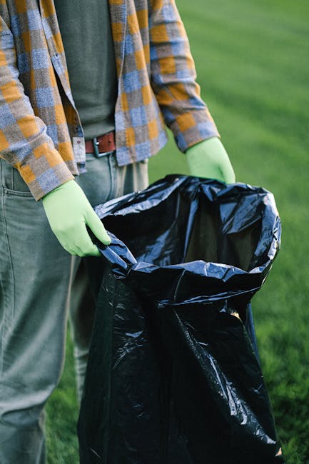 A large open-top metal bin situated on a paved urban street, overflowing with various types of waste and rubbish, including cardboard boxes, plastic bags, paper debris, and household packaging. The bi