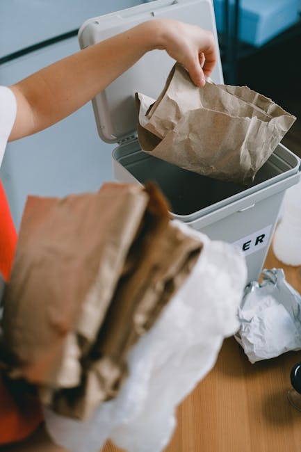 A person is seen disposing of paper waste into a grey waste bin during a rubbish removal session, with their hand holding a crumpled paper bag and slightly opening the bin lid; in the foreground, ther